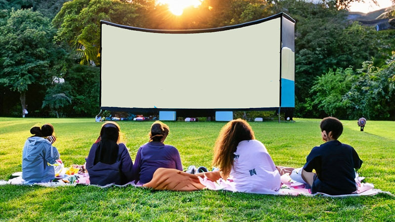 Teens sitting on a blanket placed on a green lawn getting ready to watch a movie on a large outdoor inflatable screen